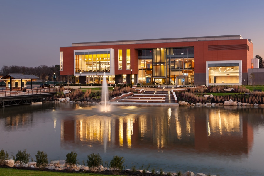 Exterior view of a modern library building at dusk, featuring large illuminated windows, a reflective pond with a fountain, and landscaped pathways, showcasing innovative and inviting design elements.