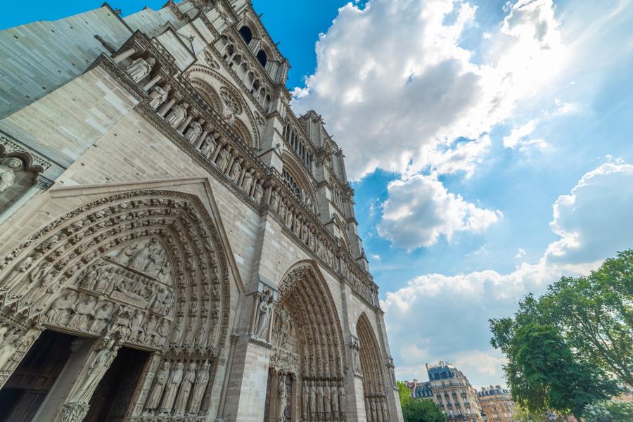 Low-angle view of the Notre-Dame Cathedral in Paris, showcasing its intricate stone carvings and Gothic architecture against a bright blue sky with fluffy clouds.