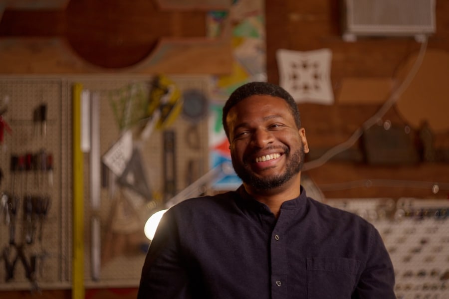 Hasheem Halim smiling in his creative workshop, surrounded by design tools and architectural elements. The African American architect and mentor is redefining architecture through community engagement and innovative design. Featured in a profile on mentorship and impact in the field.