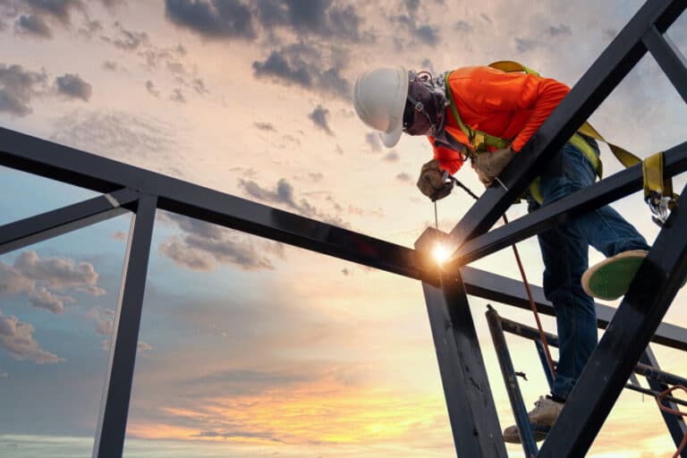 Construction worker welding steel beams at sunset, wearing safety gear and harness — symbolizing the importance of securing construction contracts amid global tariff uncertainties.