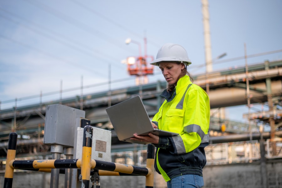 A female engineer wearing a high-visibility jacket and hard hat uses a laptop at an industrial construction site, streamlining workflows with digital tools. Learn how Plant Integrity Management improved engineering efficiency with Bluebeam.