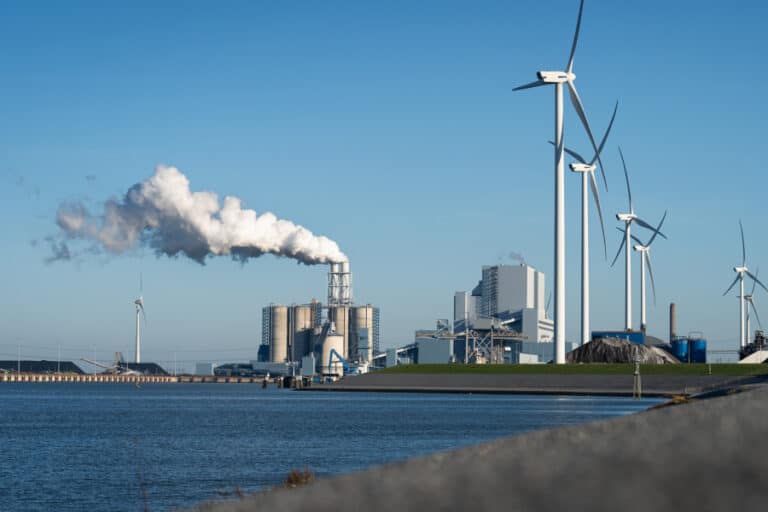 Wind turbines and oil refinery near water under blue sky—symbolizing the oil and gas industry’s transition to sustainable energy through digital collaboration.