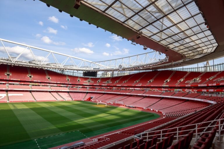 Interior view of a modern Premier League stadium with red seats, architectural roof design, and a pristine green pitch, highlighting the evolution of football stadiums into architectural icons.