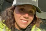 Kailyn Braddock, a female road construction worker in a reflective safety jacket and ballcap, smiles in the cab of a work truck during her night shift in Maine.