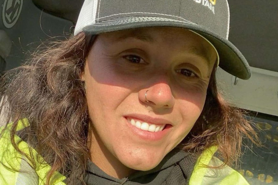Kailyn Braddock, a female road construction worker in a reflective safety jacket and ballcap, smiles in the cab of a work truck during her night shift in Maine.