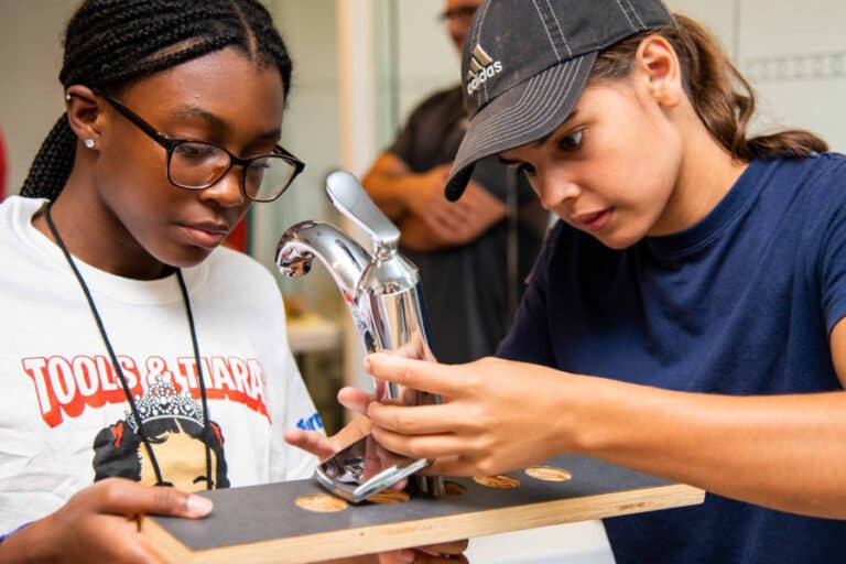 Two girls install a faucet during a Tools & Tiaras construction camp, learning hands-on plumbing skills to empower young women in the trades.