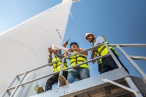Three engineers in safety vests reviewing wind turbine plans on-site, highlighting collaboration challenges in green energy construction projects.