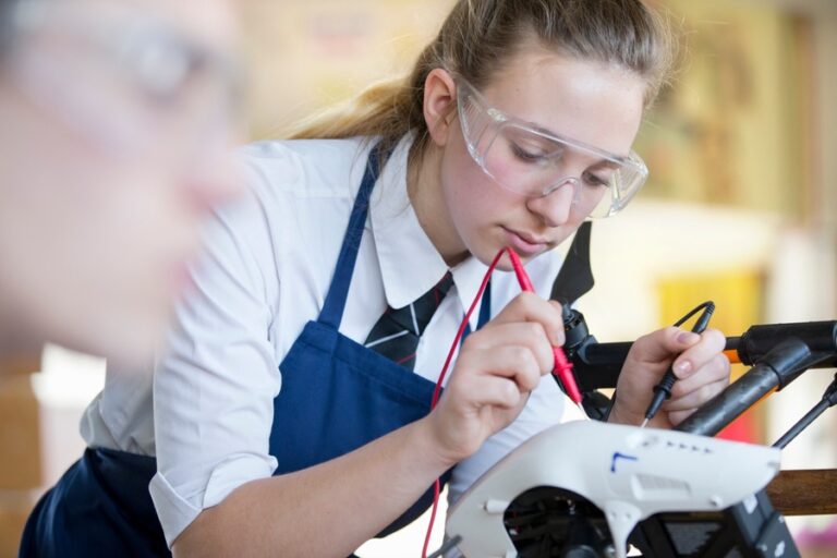 A high school girl in safety goggles uses electrical probes on a drone during shop class, symbolizing the resurgence of hands-on technical education in modern construction careers.