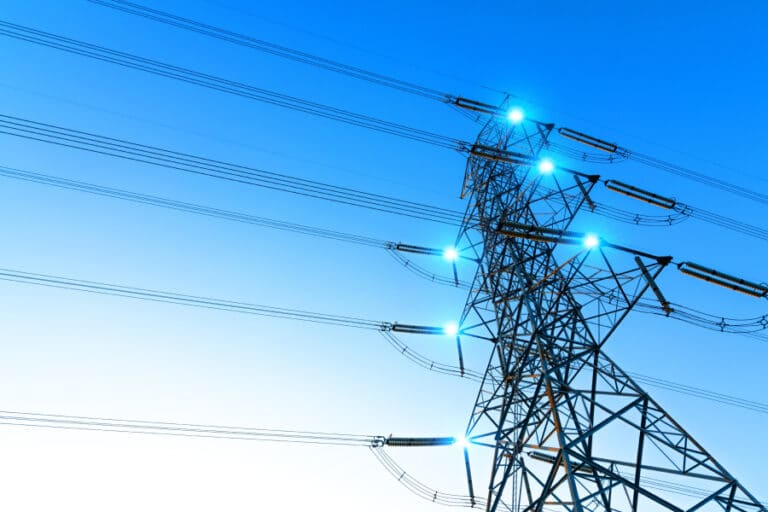 High-voltage power lines on a steel transmission tower at sunset, symbolizing the infrastructure challenges—like copper supply, concrete demand, and permitting delays—that threaten to bottleneck AI expansion.