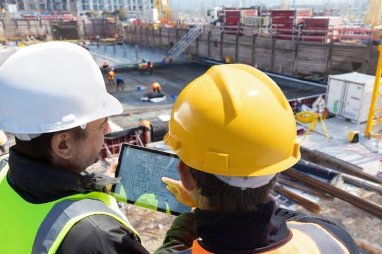 Two construction workers in safety gear review digital blueprints on a tablet at an active jobsite, demonstrating the use of integrated construction software tools to streamline project workflows.