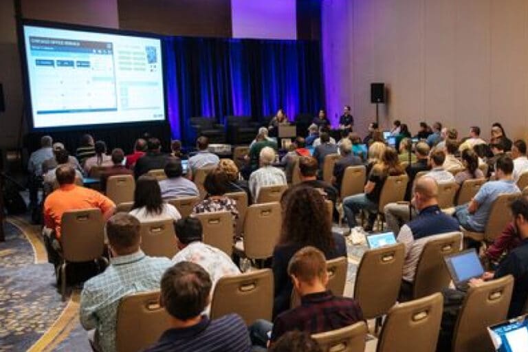 Attendees listen to a live panel session at Bluebeam's Unbound 2025 conference, with speakers presenting in front of a large screen and blue-lit stage.