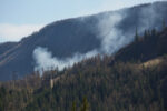 Smoke from a wildfire rises through a dense mountain forest, threatening nearby power lines and utility infrastructure—illustrating the urgent race utilities face to protect the grid from wildfire damage.
