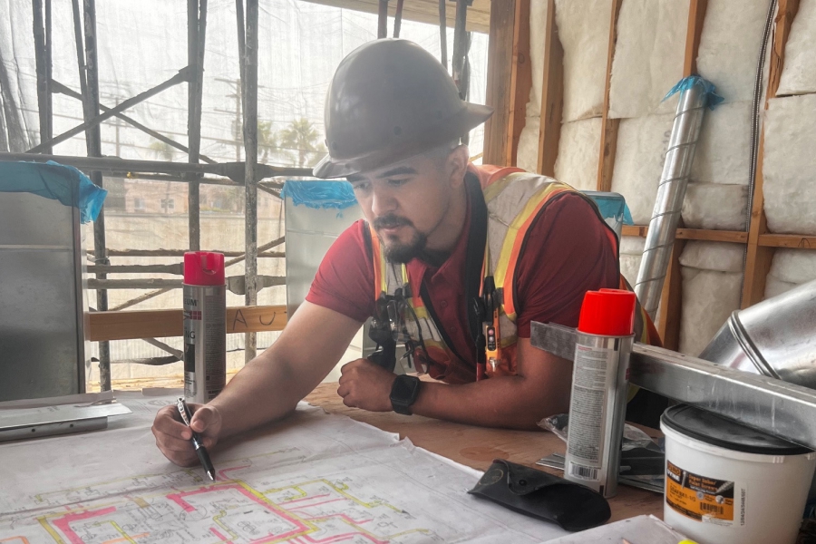 Superintendent Bob Medina reviews blueprints at a construction site, wearing a hard hat and safety vest. He marks up plans while coordinating project details, reflecting his dual role as a jobsite leader and online mentor for tradespeople.