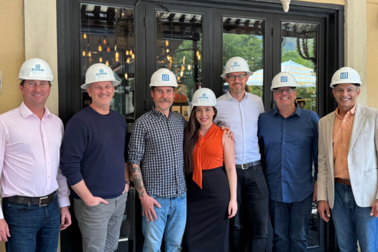 Bluebeam executive team posing with Firmus founder Shir Abecasis, all wearing Bluebeam-branded hard hats, smiling in front of a restaurant patio entrance during a construction industry partnership event.