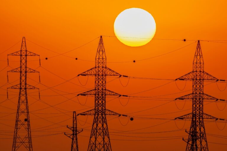 High-voltage power transmission towers silhouetted against the sun, illustrating how AI-driven demand is stressing U.S. power grid infrastructure and exposing permitting delays.
