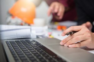 Estimator reviewing revised construction drawings and performing digital takeoff on laptop in jobsite trailer during project revision.