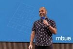 Jean-Pierre Trou, founder of mbue, presenting on stage with a construction drawing blueprint overlay on a Bluebeam blue background.