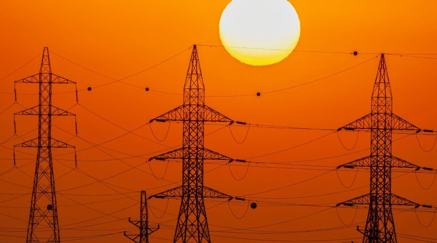High-voltage power transmission towers silhouetted against the sun, illustrating how AI-driven demand is stressing U.S. power grid infrastructure and exposing permitting delays.