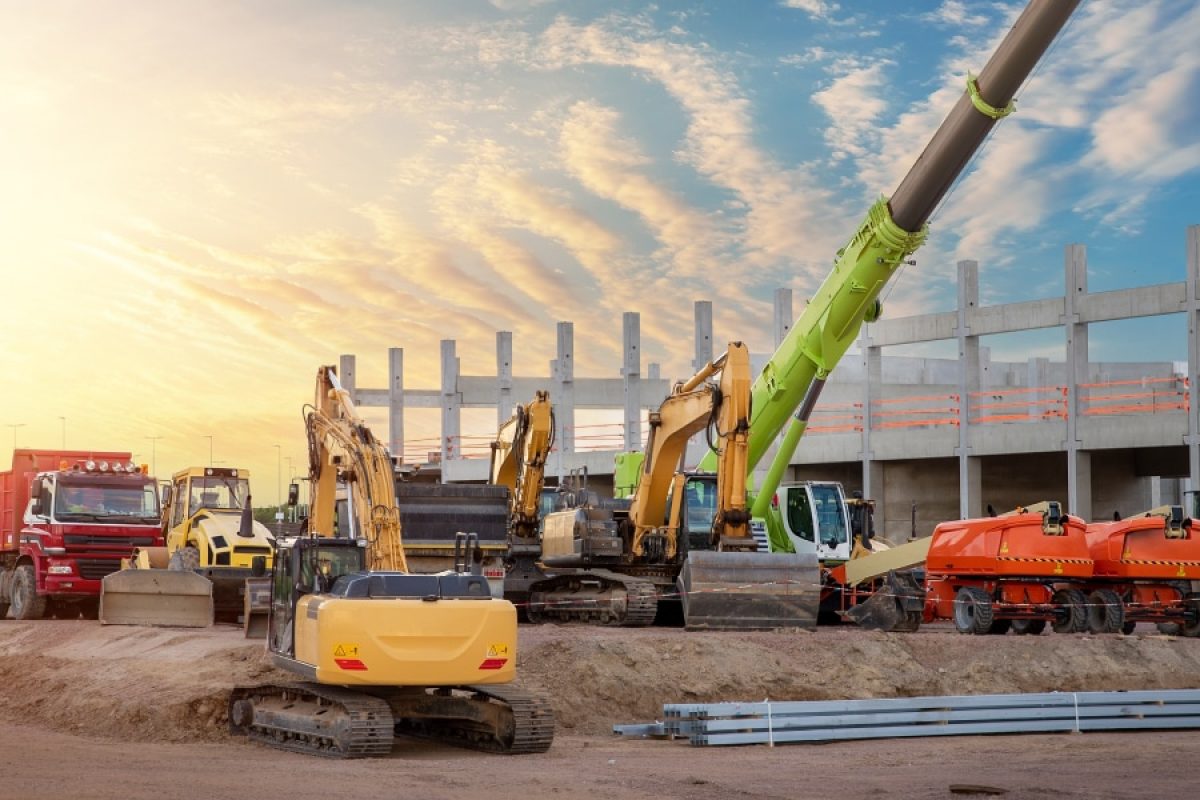 The image depicts a construction site at sunset, featuring various heavy machinery, including excavators, a crane, and trucks, positioned around a partially built concrete structure. The sky is vibrant with warm hues, emphasizing the contrast between the industrial scene and the natural backdrop.