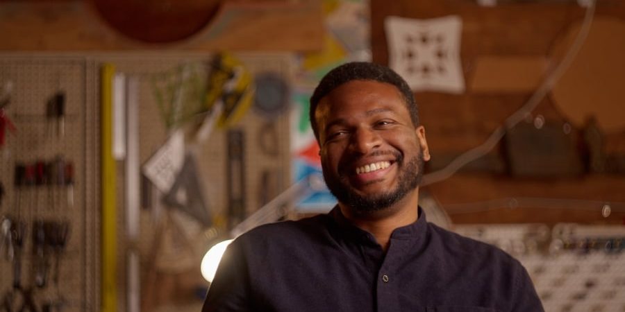 Hasheem Halim smiling in his creative workshop, surrounded by design tools and architectural elements. The African American architect and mentor is redefining architecture through community engagement and innovative design. Featured in a profile on mentorship and impact in the field.