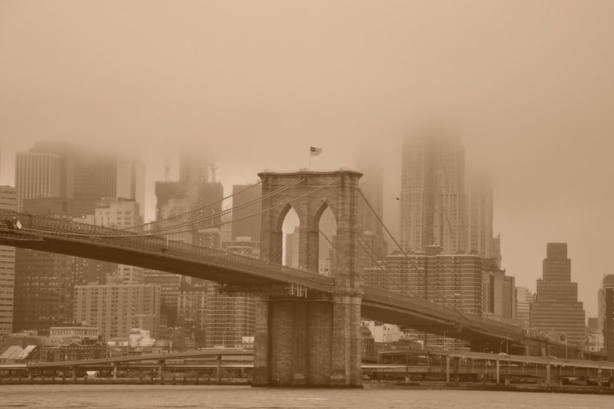 Sepia-toned image of the Brooklyn Bridge enveloped in fog, with the New York City skyline in the background. The bridge, completed under the leadership of Emily Warren Roebling, stands as a testament to her pivotal role in its construction.