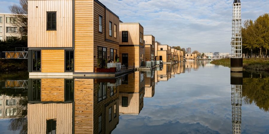 Modern floating homes with sustainable wooden exteriors reflecting on calm water in an urban canal, showcasing innovative floating architecture as a solution to urban space challenges.