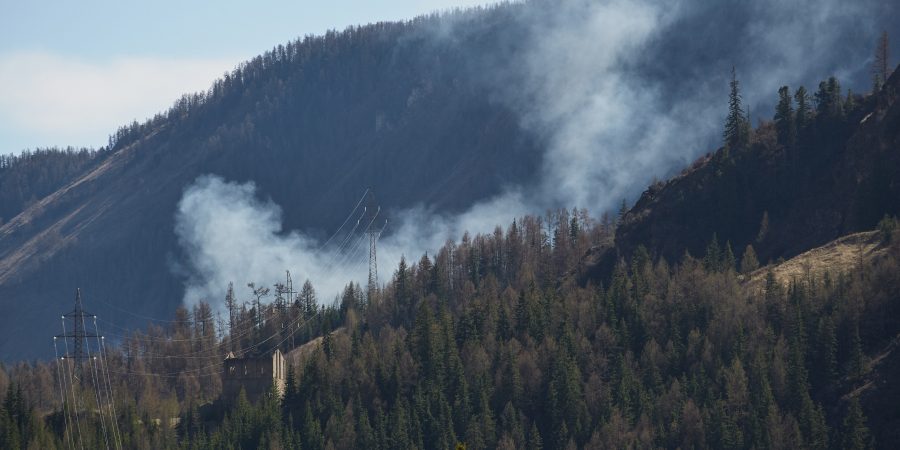 Smoke from a wildfire rises through a dense mountain forest, threatening nearby power lines and utility infrastructure—illustrating the urgent race utilities face to protect the grid from wildfire damage.