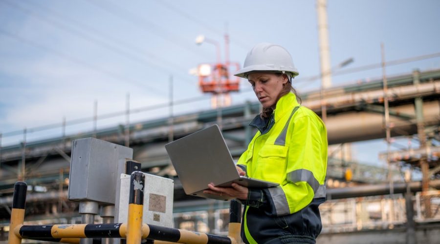 A female engineer wearing a high-visibility jacket and hard hat uses a laptop at an industrial construction site, streamlining workflows with digital tools. Learn how Plant Integrity Management improved engineering efficiency with Bluebeam.