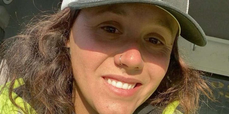Kailyn Braddock, a female road construction worker in a reflective safety jacket and ballcap, smiles in the cab of a work truck during her night shift in Maine.