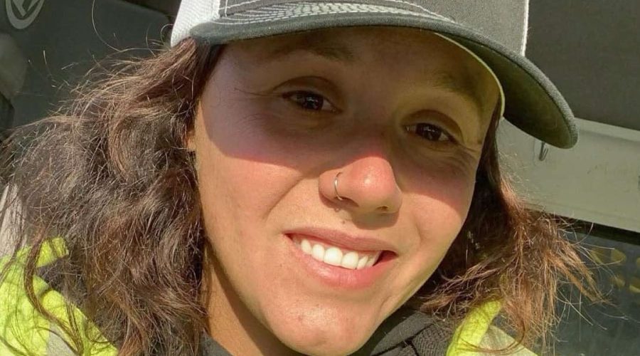 Kailyn Braddock, a female road construction worker in a reflective safety jacket and ballcap, smiles in the cab of a work truck during her night shift in Maine.
