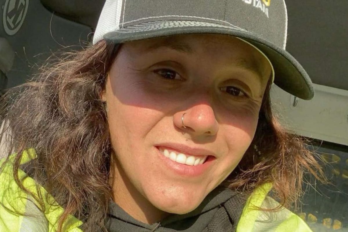 Kailyn Braddock, a female road construction worker in a reflective safety jacket and ballcap, smiles in the cab of a work truck during her night shift in Maine.