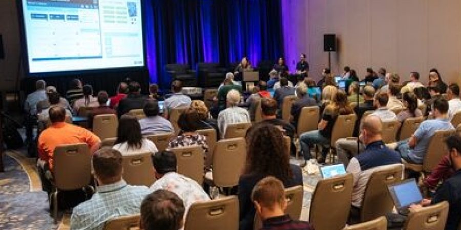 Attendees listen to a live panel session at Bluebeam's Unbound 2025 conference, with speakers presenting in front of a large screen and blue-lit stage.