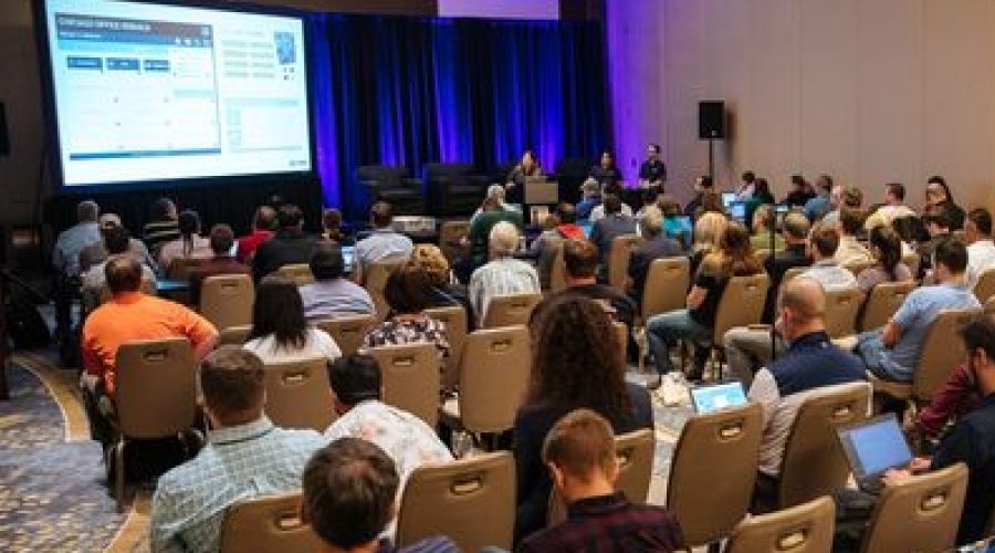 Attendees listen to a live panel session at Bluebeam's Unbound 2025 conference, with speakers presenting in front of a large screen and blue-lit stage.