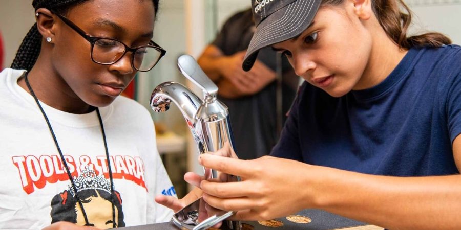 Two girls install a faucet during a Tools & Tiaras construction camp, learning hands-on plumbing skills to empower young women in the trades.