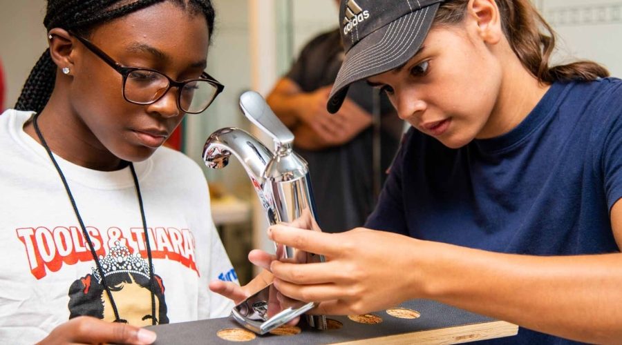 Two girls install a faucet during a Tools & Tiaras construction camp, learning hands-on plumbing skills to empower young women in the trades.