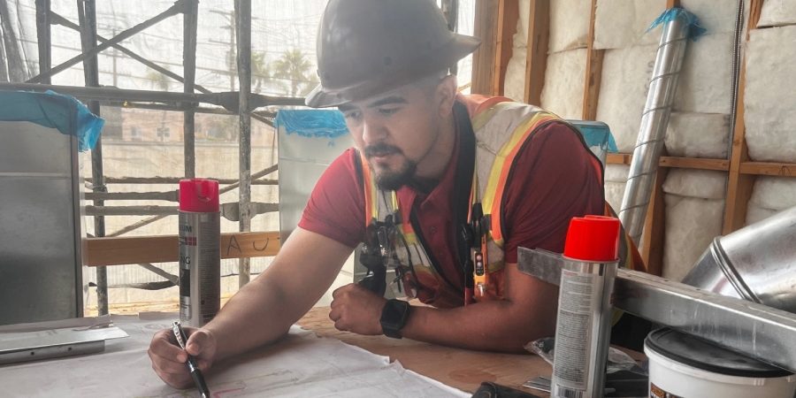 Superintendent Bob Medina reviews blueprints at a construction site, wearing a hard hat and safety vest. He marks up plans while coordinating project details, reflecting his dual role as a jobsite leader and online mentor for tradespeople.