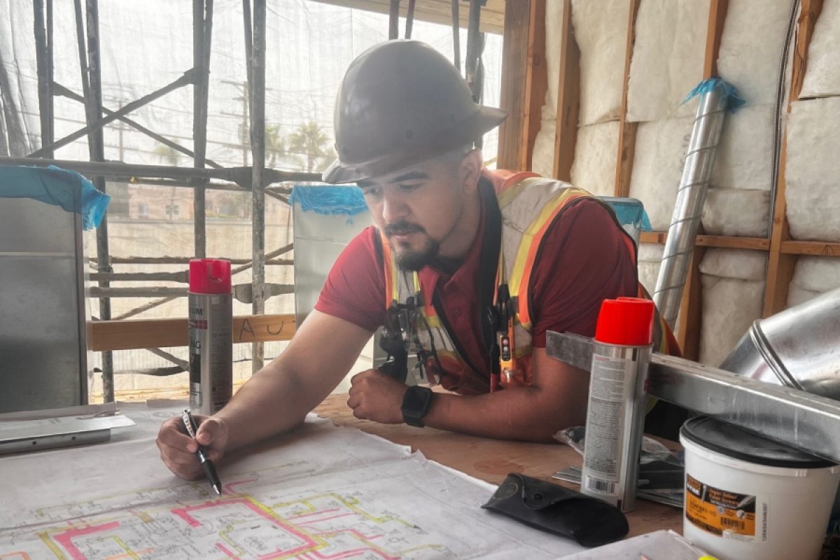 Superintendent Bob Medina reviews blueprints at a construction site, wearing a hard hat and safety vest. He marks up plans while coordinating project details, reflecting his dual role as a jobsite leader and online mentor for tradespeople.