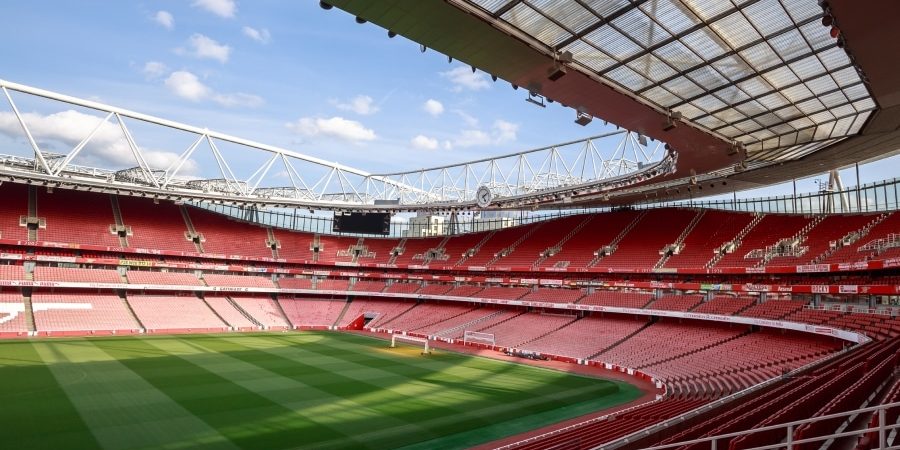 Interior view of a modern Premier League stadium with red seats, architectural roof design, and a pristine green pitch, highlighting the evolution of football stadiums into architectural icons.