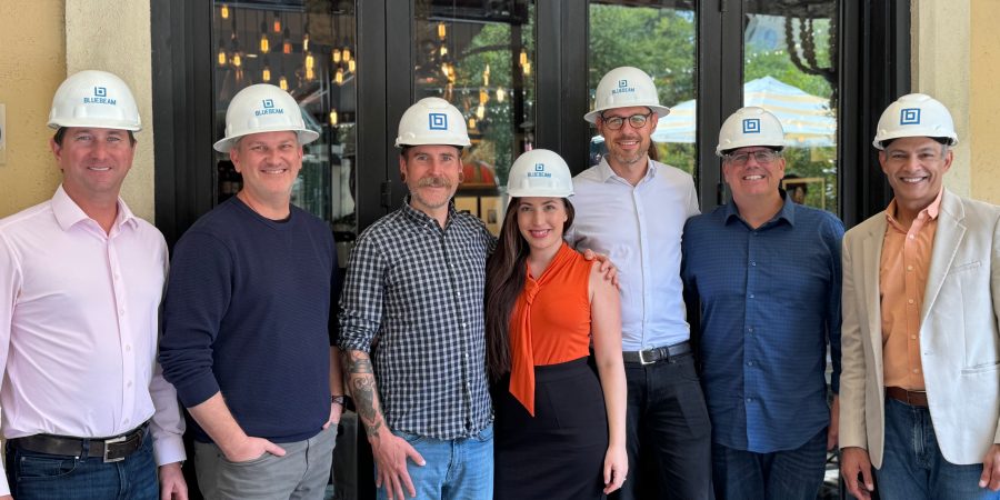 Bluebeam executive team posing with Firmus founder Shir Abecasis, all wearing Bluebeam-branded hard hats, smiling in front of a restaurant patio entrance during a construction industry partnership event.