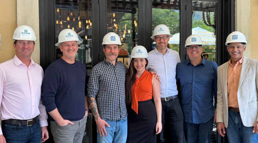 Bluebeam executive team posing with Firmus founder Shir Abecasis, all wearing Bluebeam-branded hard hats, smiling in front of a restaurant patio entrance during a construction industry partnership event.
