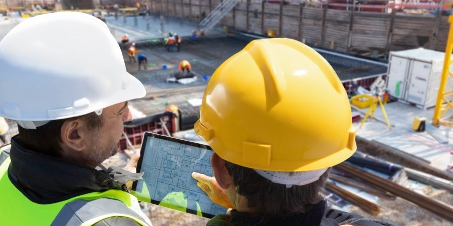 Two construction workers in safety gear review digital blueprints on a tablet at an active jobsite, demonstrating the use of integrated construction software tools to streamline project workflows.