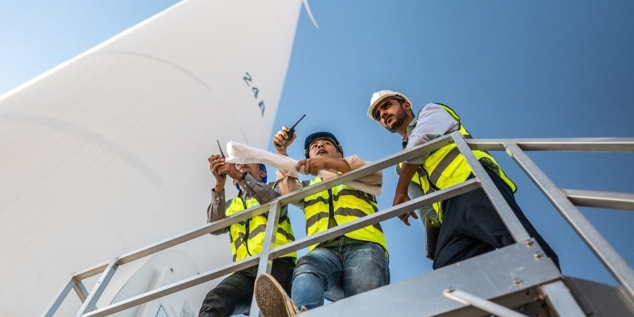 Three engineers in safety vests reviewing wind turbine plans on-site, highlighting collaboration challenges in green energy construction projects.
