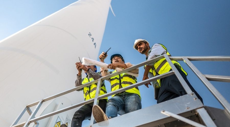Three engineers in safety vests reviewing wind turbine plans on-site, highlighting collaboration challenges in green energy construction projects.