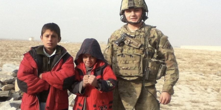 Josh Sergent, in full U.S. Air Force combat gear, stands with two local Afghan children wearing red jackets during a patrol mission, symbolizing his service abroad before transitioning into a career in construction and customer success at Bluebeam.