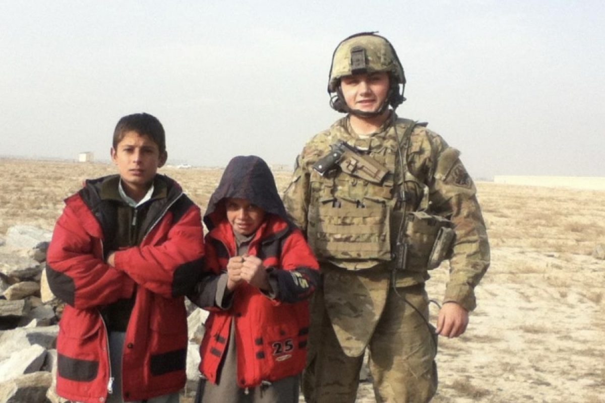 Josh Sergent, in full U.S. Air Force combat gear, stands with two local Afghan children wearing red jackets during a patrol mission, symbolizing his service abroad before transitioning into a career in construction and customer success at Bluebeam.