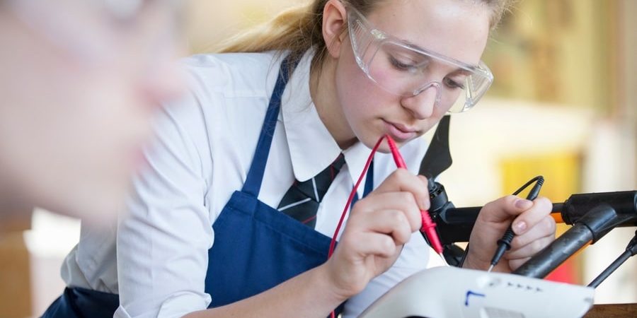 A high school girl in safety goggles uses electrical probes on a drone during shop class, symbolizing the resurgence of hands-on technical education in modern construction careers.