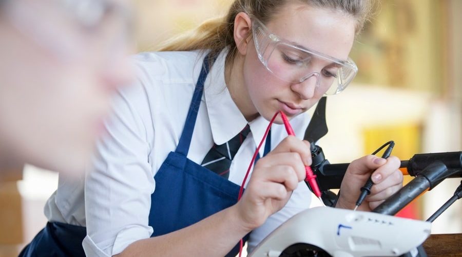A high school girl in safety goggles uses electrical probes on a drone during shop class, symbolizing the resurgence of hands-on technical education in modern construction careers.
