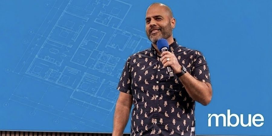 Jean-Pierre Trou, founder of mbue, presenting on stage with a construction drawing blueprint overlay on a Bluebeam blue background.