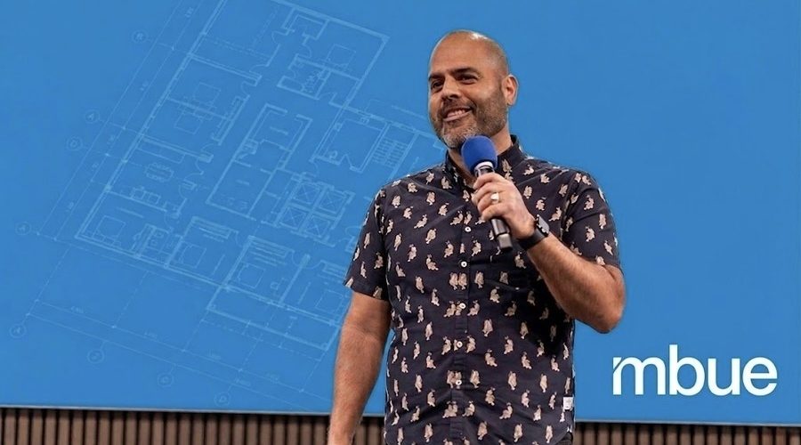 Jean-Pierre Trou, founder of mbue, presenting on stage with a construction drawing blueprint overlay on a Bluebeam blue background.