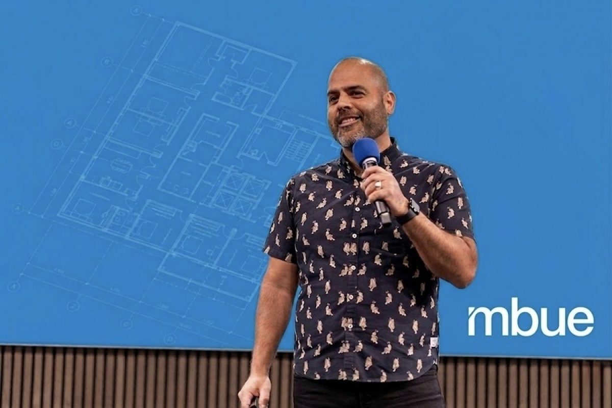 Jean-Pierre Trou, founder of mbue, presenting on stage with a construction drawing blueprint overlay on a Bluebeam blue background.